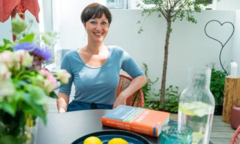 Dr. Julia Belke – Online-Psychotherapie in entspannter Atmosphäre Psychotherapeutin Julia Belke sitzt auf der Terrasse mit einem Buch über Selbstfürsorge, Zitrone-Wasser und Blumen auf dem Tisch"